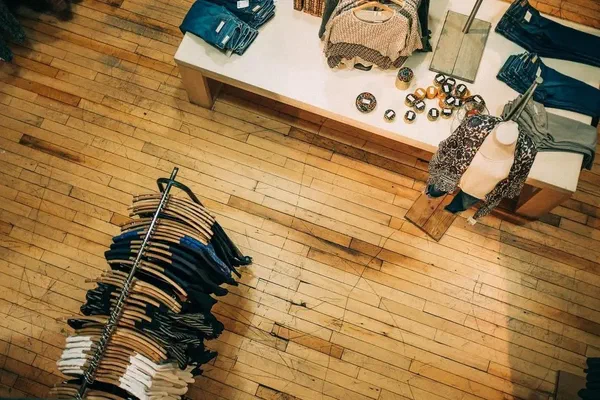 A retail store with clothing racks, a display table, and wood floors. This retail store is looking bland and is in need of digital store signs.
