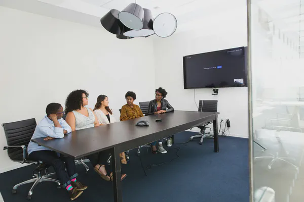 A meeting room filled with employees around a large table and a meeting room digital sign on the wall behind them.